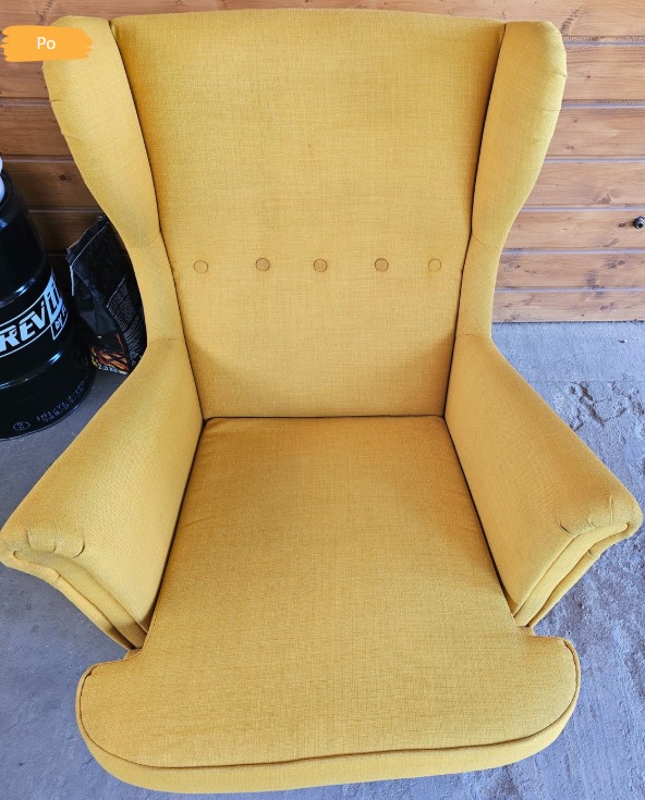 Żółty fotel typu uszak po praniu tapicerki, z widoczną etykietą 'Po' w rogu, na tle drewnianej ściany i betonowej podłogi.