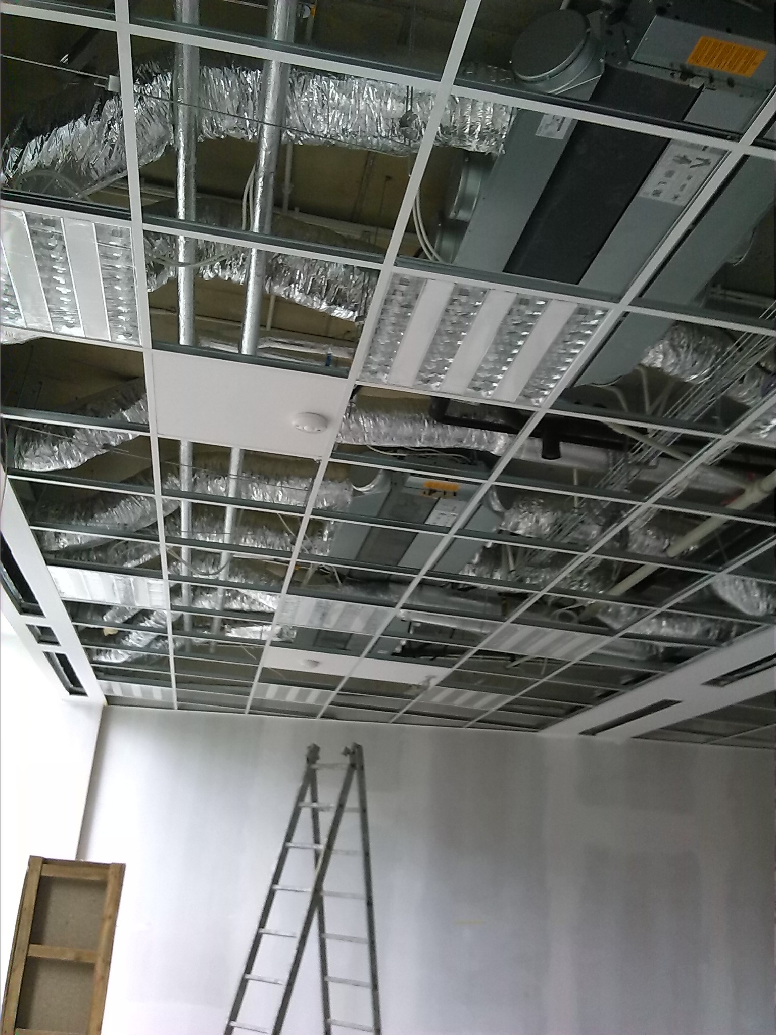 Widok sufitu podwieszanego w trakcie montażu, widoczne elementy konstrukcyjne, instalacje wentylacyjne w srebrnej izolacji, oświetlenie, drabina oparta o ścianę i fragmenty niezabudowanej...