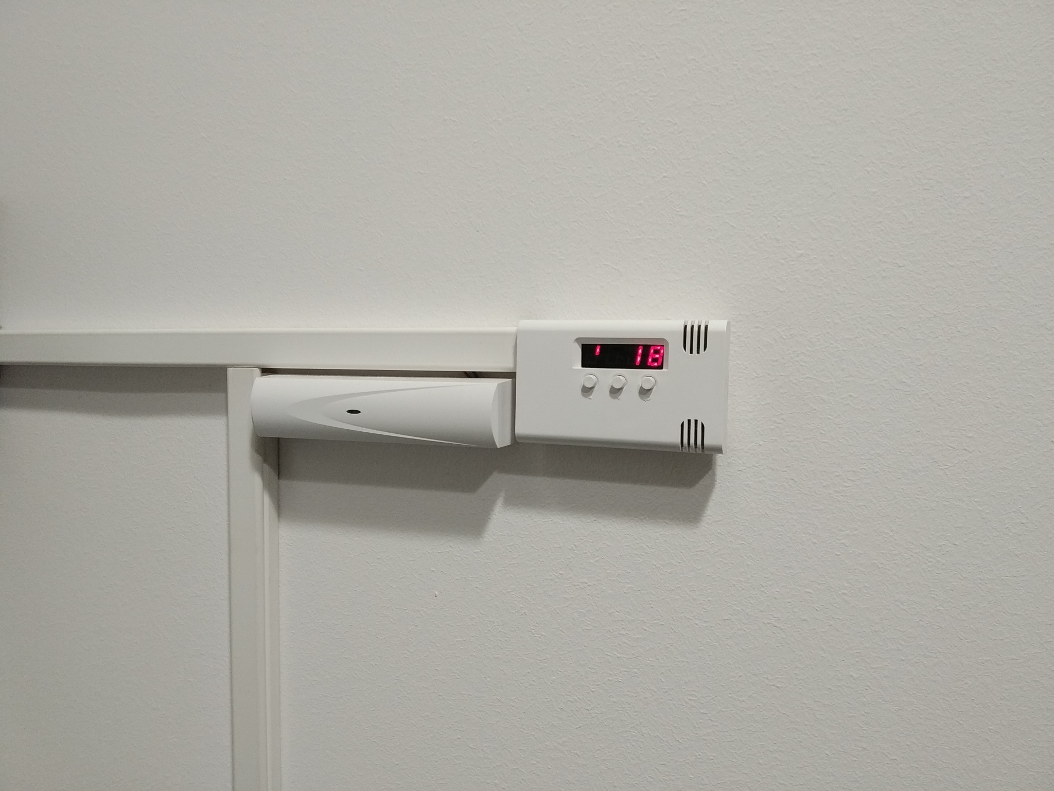 Biały czujnik alarmowy z wyświetlaczem LED i trzema przyciskami, zamontowany na ścianie, widoczny fragment listwy przypodłogowej.