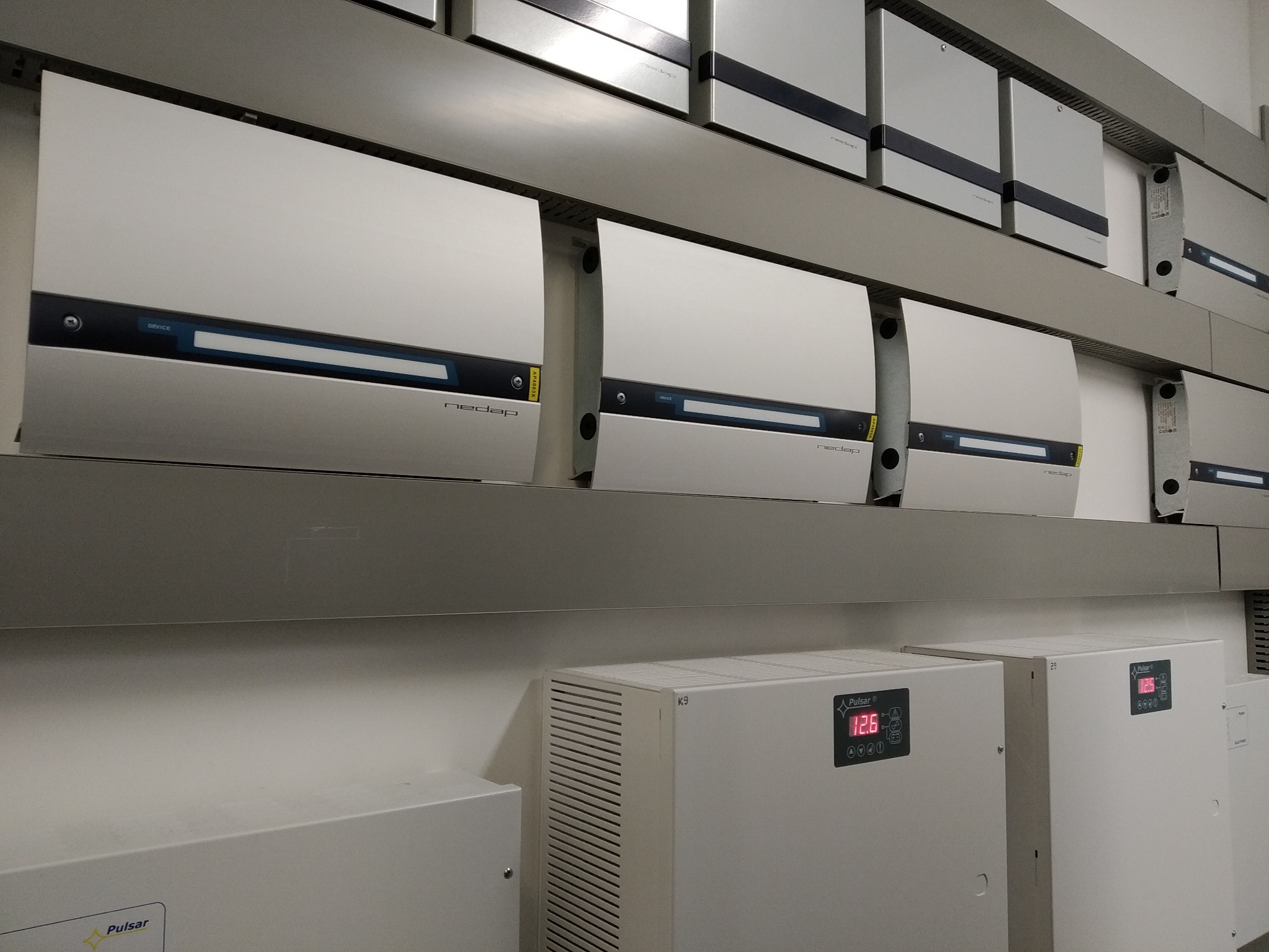 Rząd białych urządzeń Nedap AP4003X zamontowanych na metalowej półce, z widocznymi panelami sterowania i napisami, poniżej dwa urządzenia Pulsar z wyświetlaczami LED wskazującymi wartości 12.6...
