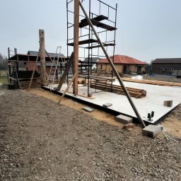 KDEM - Wznoszenie konstrukcji drewnianej budynku: drewniane słupy wsparte na betonowej płycie fundamentowej, metalowe rusztowanie, przygotowane belki, żwirowe podłoże.