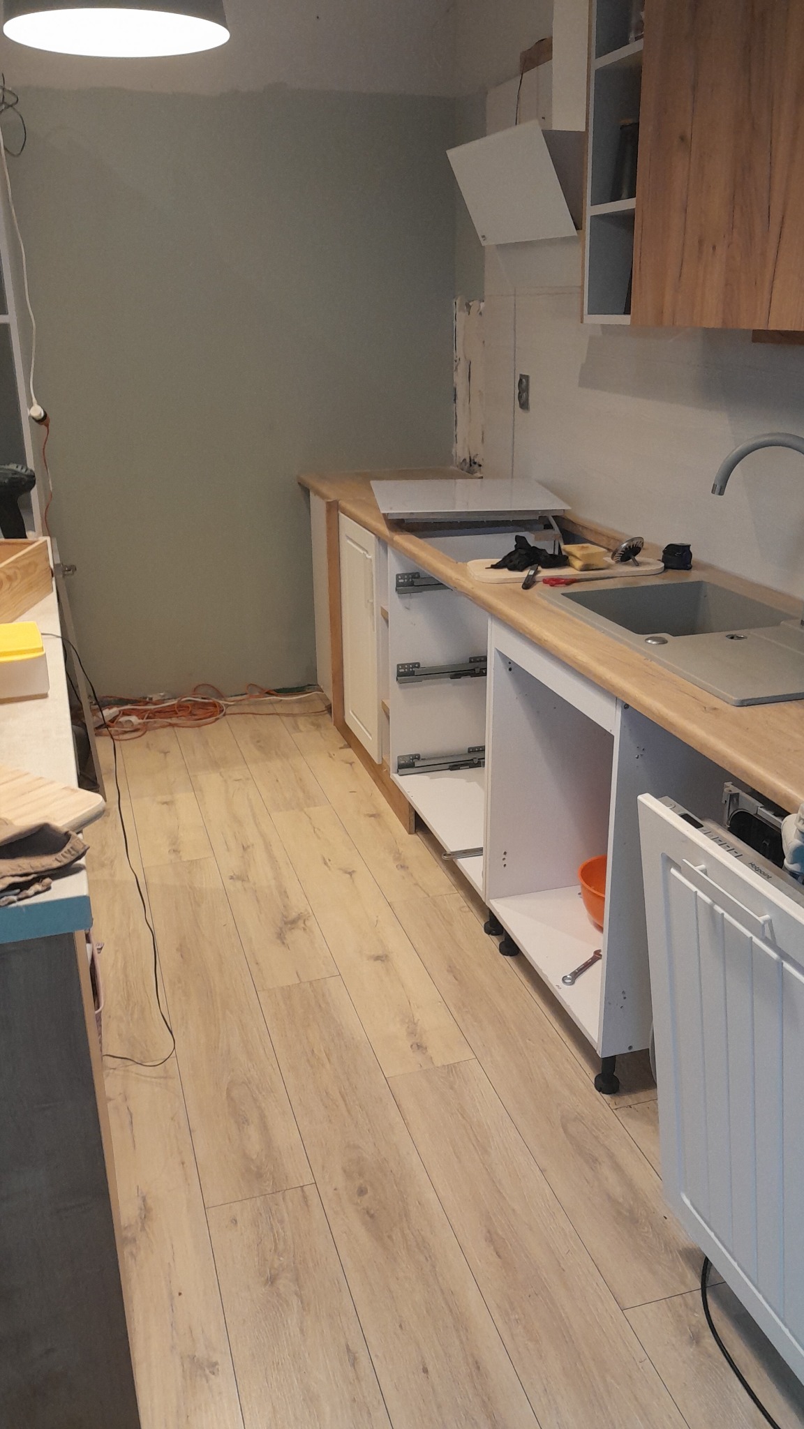 Montaż szafek kuchennych z drewnianym blatem i szarą umywalką, widoczne niedokończone podłączenia, narzędzia na blacie i pomarańczowe kable na podłodze.