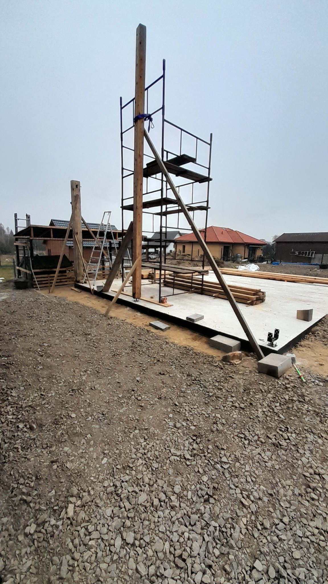 Wznoszenie konstrukcji drewnianej budynku: drewniane słupy wsparte na betonowej płycie fundamentowej, metalowe rusztowanie, przygotowane belki, żwirowe podłoże.