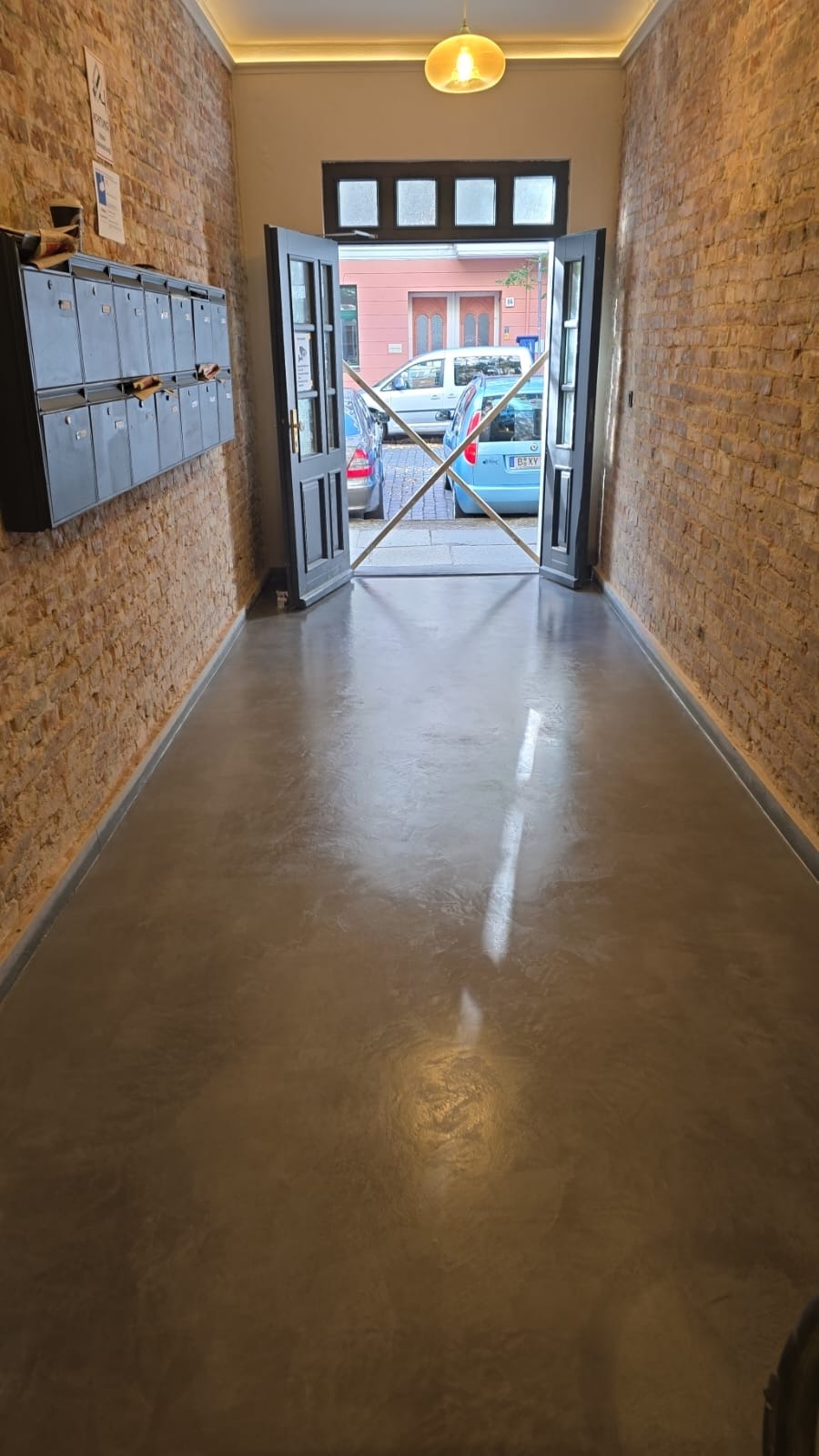 Wnętrze korytarza z podłogą mikrocementową w odcieniach szarości i brązu, ściany z cegły, skrzynki pocztowe po lewej stronie, otwarte drzwi wejściowe.