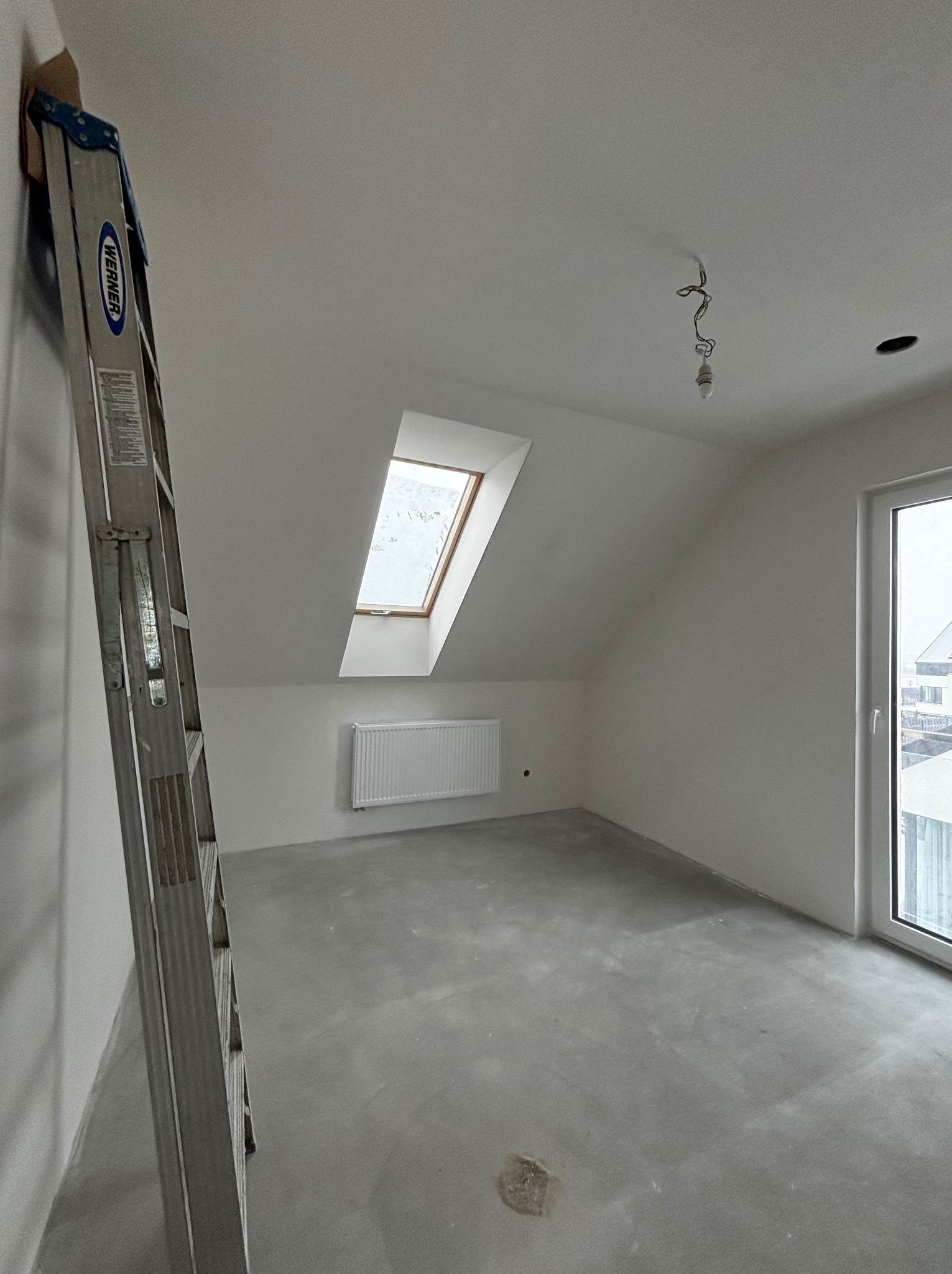 Pokój na poddaszu ze skośnym oknem dachowym, grzejnikiem, szarą wylewką i rozłożoną drabiną Werner przy ścianie, przygotowany do dalszych prac wykończeniowych.