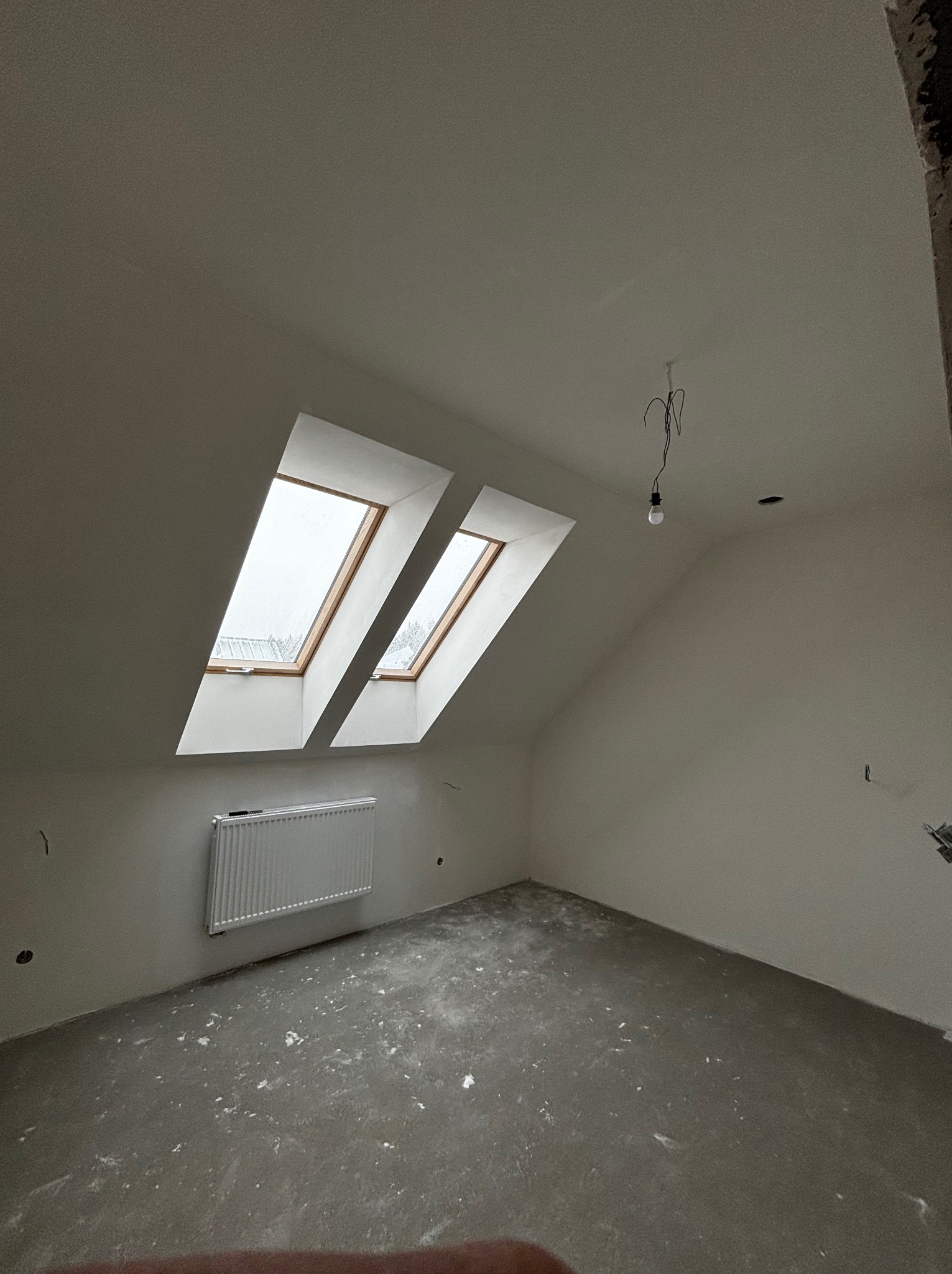 Poddasze w trakcie prac wykończeniowych: widoczne dwa okna dachowe, grzejnik na ścianie, surowy beton na podłodze, wisząca żarówka i ślady po instalacjach elektrycznych.