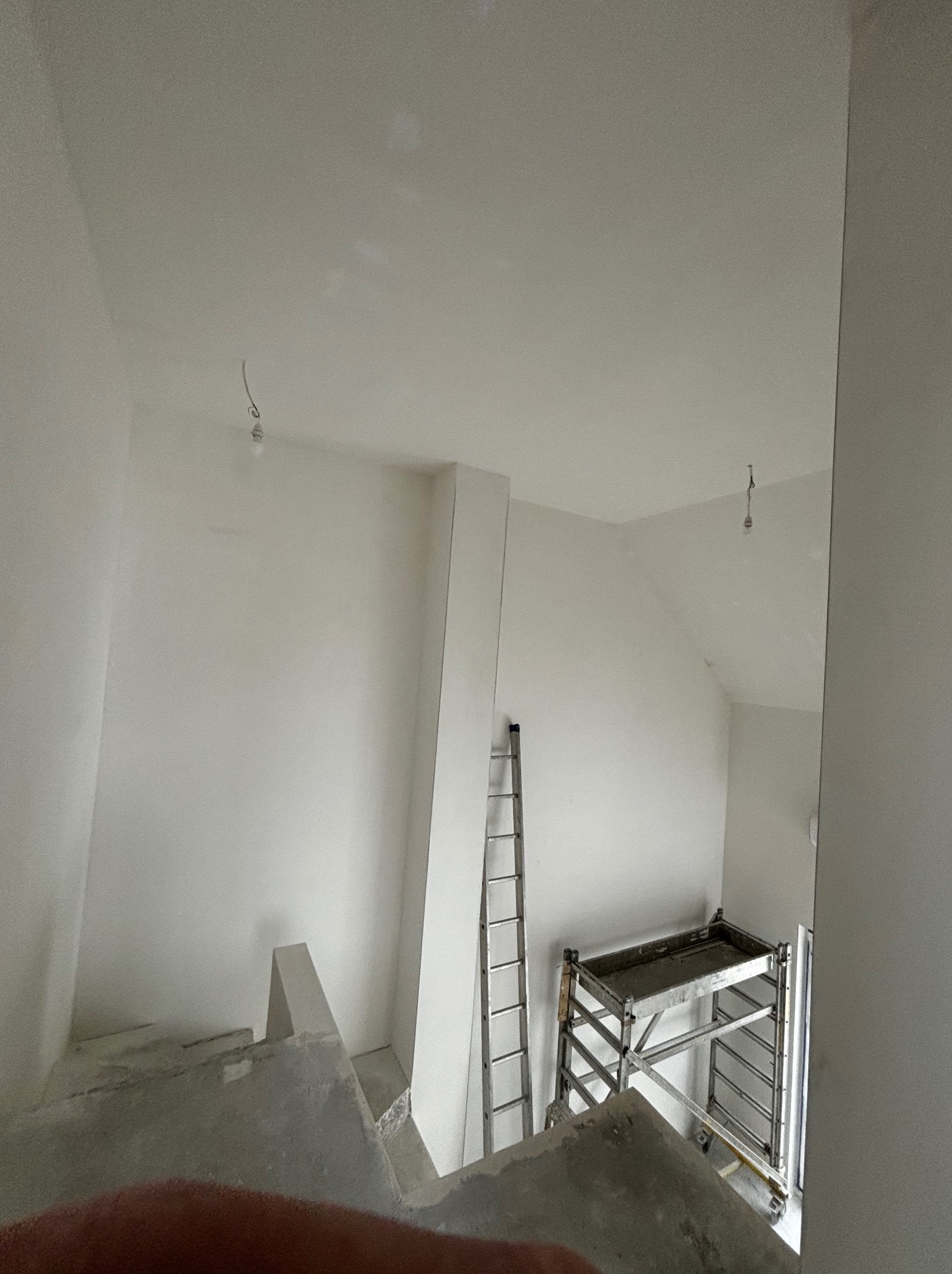 Wnętrze w trakcie wykańczania: pomalowane na biało ściany, aluminiowa drabina oparta o ścianę, metalowe rusztowanie, surowy beton na schodach, goła żarówka na suficie.