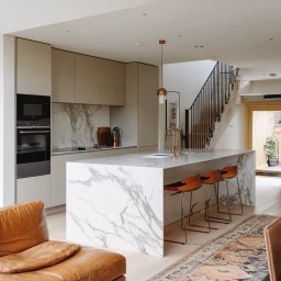Kasus Studio - Nowoczesna kuchnia z marmurowym blatem wyspowym, drewnianymi krzesłami barowymi i beżowymi szafkami, widok na schody z czarną balustradą.