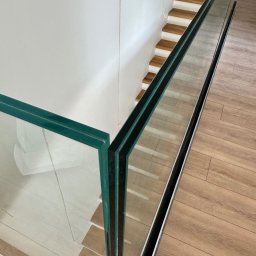 Monika TARKA - Szklana balustrada z zielonkawym odcieniem szkła, zamontowana przy schodach z drewnianymi stopniami. Widok z góry, ukazujący detale mocowania i odbicie podłogi.