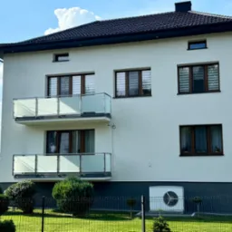 Nowoczesny dom z białym tynkiem i dwoma balkonami z szklanymi balustradami. Widoczne okna z roletami, zadbany trawnik i metalowe ogrodzenie.