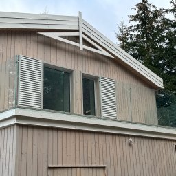 Monika TARKA - Nowoczesny balkon z balustradą szklaną, drewniane wykończenie elewacji, widok z dołu. Balustrada z elementami metalowymi i szkłem. Tło stanowią drzewa.