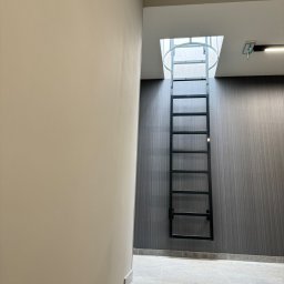 Monika TARKA - Metalowa drabina ewakuacyjna, przytwierdzona do ściany o prążkowanej strukturze, prowadząca do otworu w suficie z widocznym oświetleniem naturalnym.