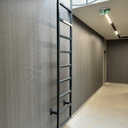 Monika TARKA - Metalowa drabina przymocowana do ściany w korytarzu, prowadząca do okrągłego otworu w suficie. Widoczny znak ewakuacyjny i oświetlenie.