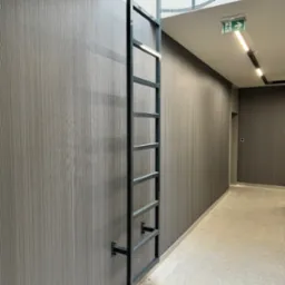 Metalowa drabina przymocowana do ściany w korytarzu, prowadząca do okrągłego otworu w suficie. Widoczny znak ewakuacyjny i oświetlenie.