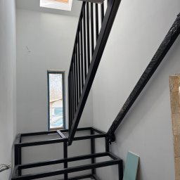 Monika TARKA - Wnętrze z metalową konstrukcją schodów z czarną balustradą. Widoczne okno i fragment osoby. Konstrukcja w trakcie montażu, zabezpieczona folią w górnej części.