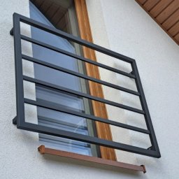 Monika TARKA - Czarna, metalowa krata okienna na elewacji budynku z oknem o drewnianych ramach. Minimalistyczny design, ochrona przed włamaniem, widok z zewnątrz.