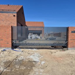 Monika TARKA - Nowoczesna brama wjazdowa z pionowych, szarych profili stalowych, wkomponowana w mur z czerwonej cegły na tle domu z czerwoną dachówką w słoneczny dzień.