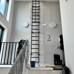 Monika TARKA - Wnętrze z metalową drabiną z obręczami ochronnymi prowadzącą do świetlika. Na schodach narzędzia i zabezpieczona folią balustrada. Widoczny fragment barierki i numer '2' na ścianie.