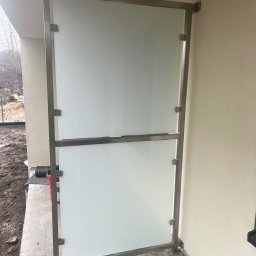 Monika TARKA - Szklana balustrada balkonowa z metalową ramą, zamontowana na betonowej posadzce. Widoczne mocowania i fragment elewacji budynku w neutralnym odcieniu.