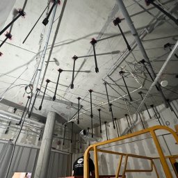 Monika TARKA - Montaż instalacji podwieszanej pod stropem, widoczne elementy mocujące i rury, w tle fragment podnośnika. Wnętrze w stanie surowym.