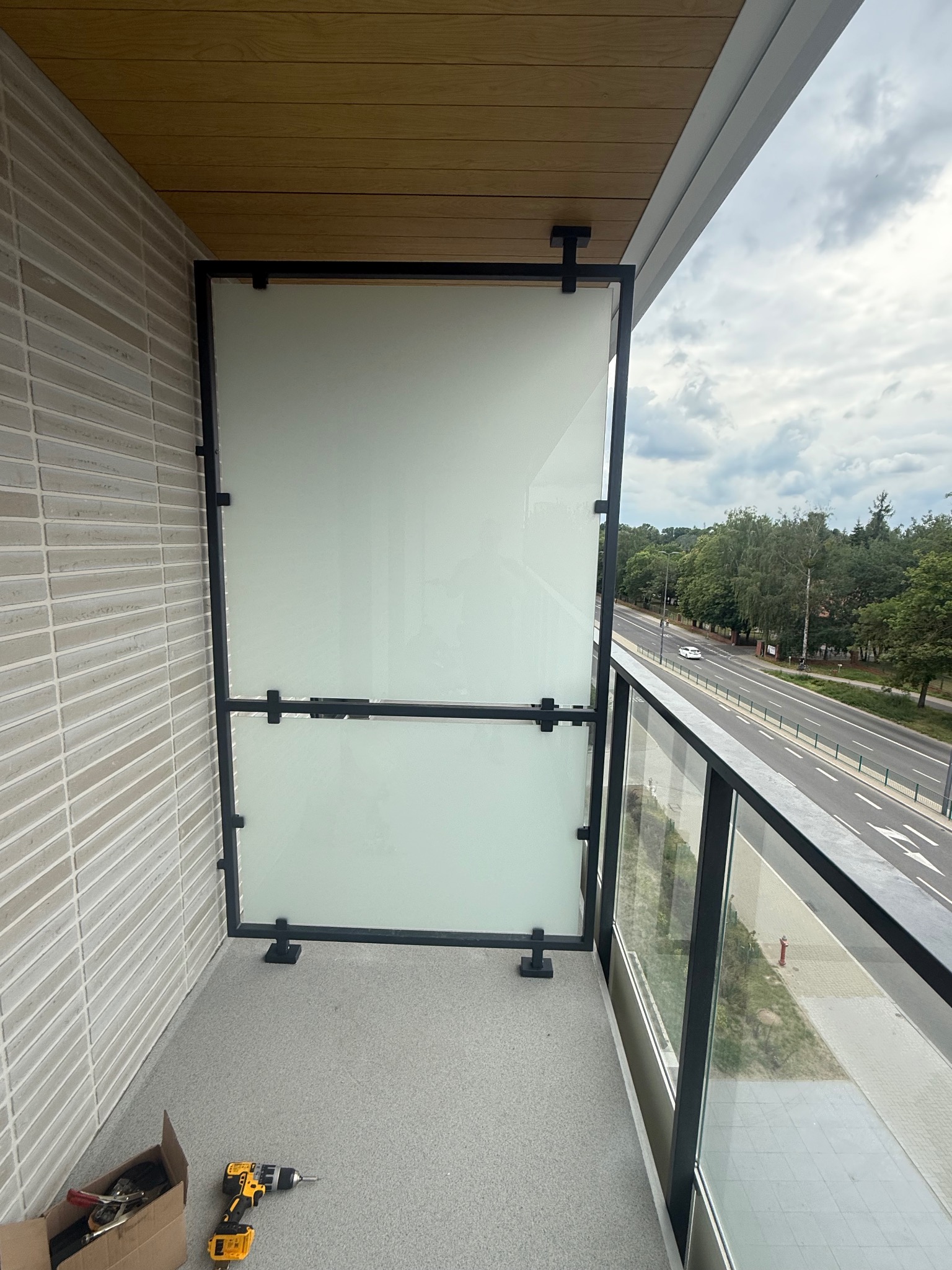 Szklana przegroda balkonowa z czarną ramą, widok na ulicę. Widoczne narzędzia pracy: wiertarka i karton z akcesoriami, płytki na ścianie balkonu.