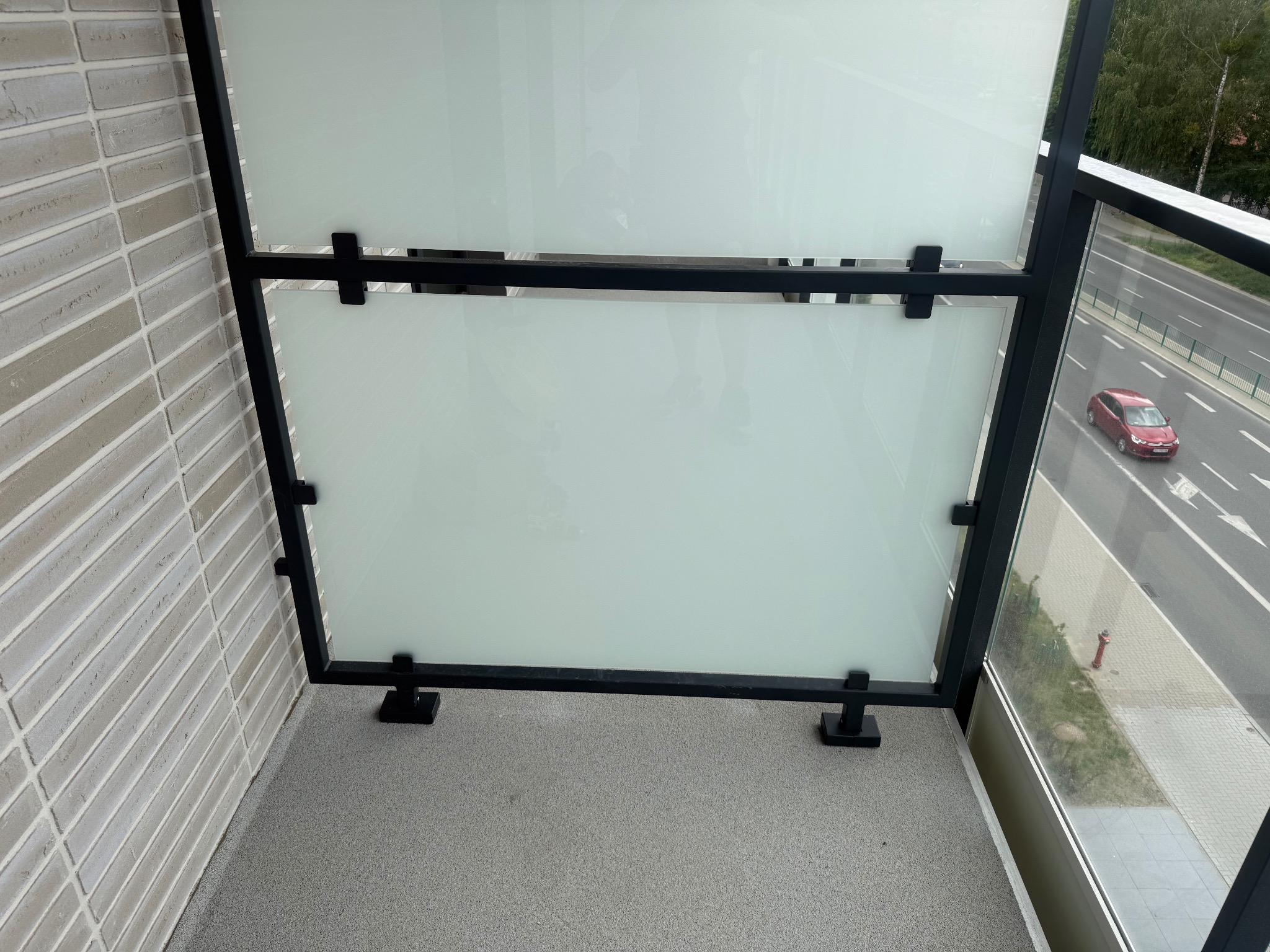 Szklana balustrada balkonowa z czarną metalową ramą, widok z góry na szary balkon i ulicę z czerwonym samochodem.