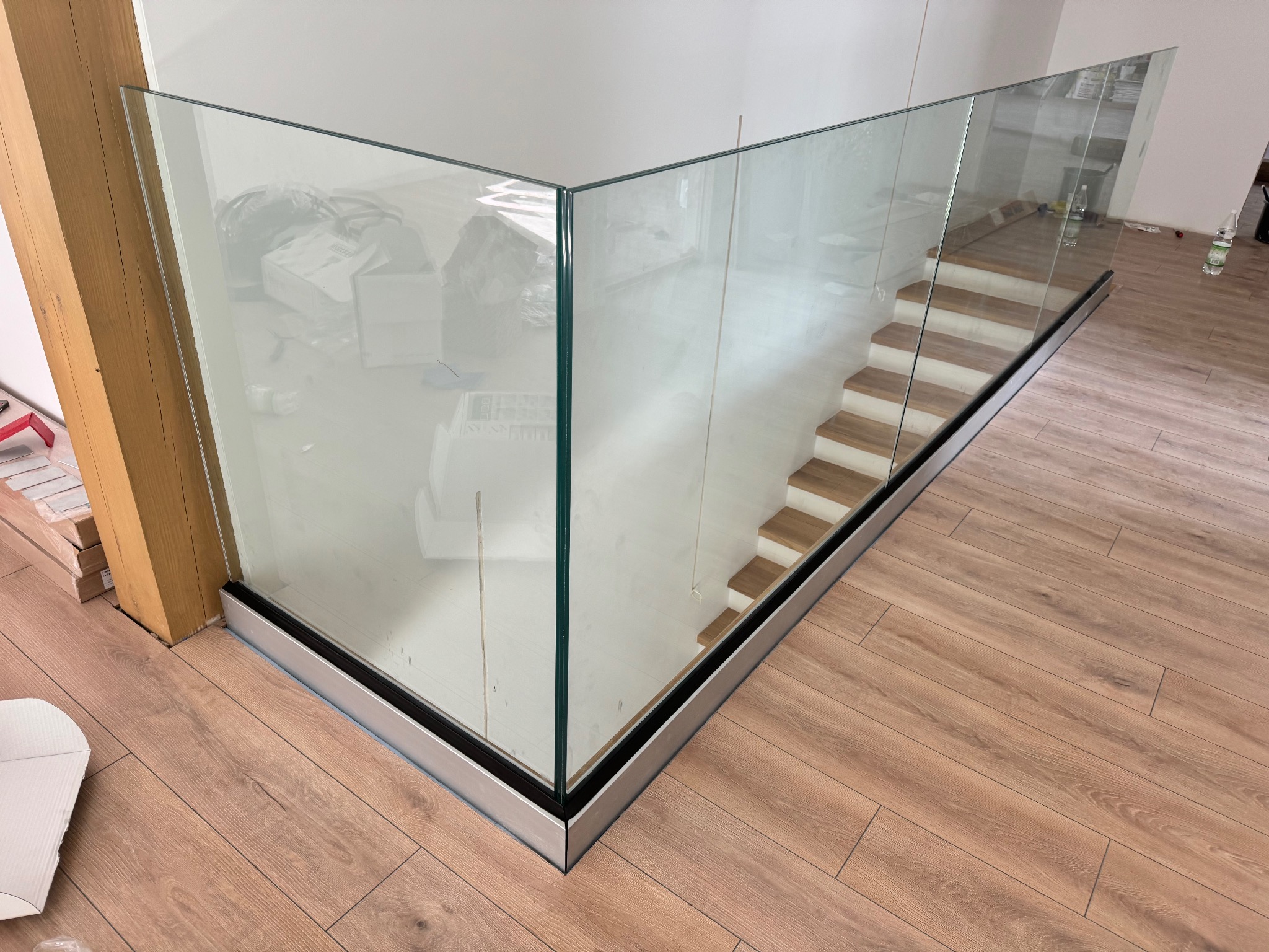 Szklana balustrada z metalową podstawą, zabezpieczająca schody z drewnianymi stopniami. Widok z góry na drewnianą podłogę i fragment pomieszczenia w tle.