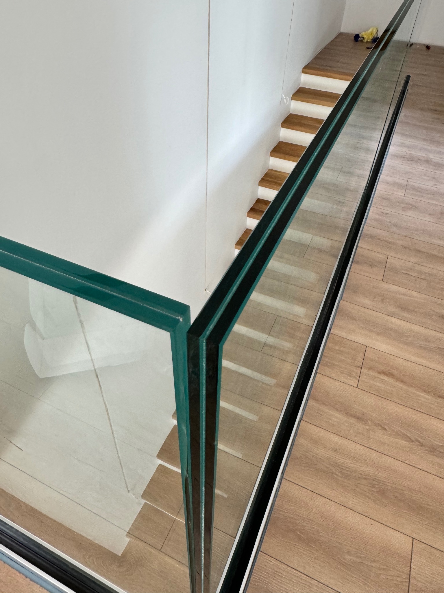 Szklana balustrada z zielonkawym odcieniem szkła, zamontowana przy schodach z drewnianymi stopniami. Widok z góry, ukazujący detale mocowania i odbicie podłogi.