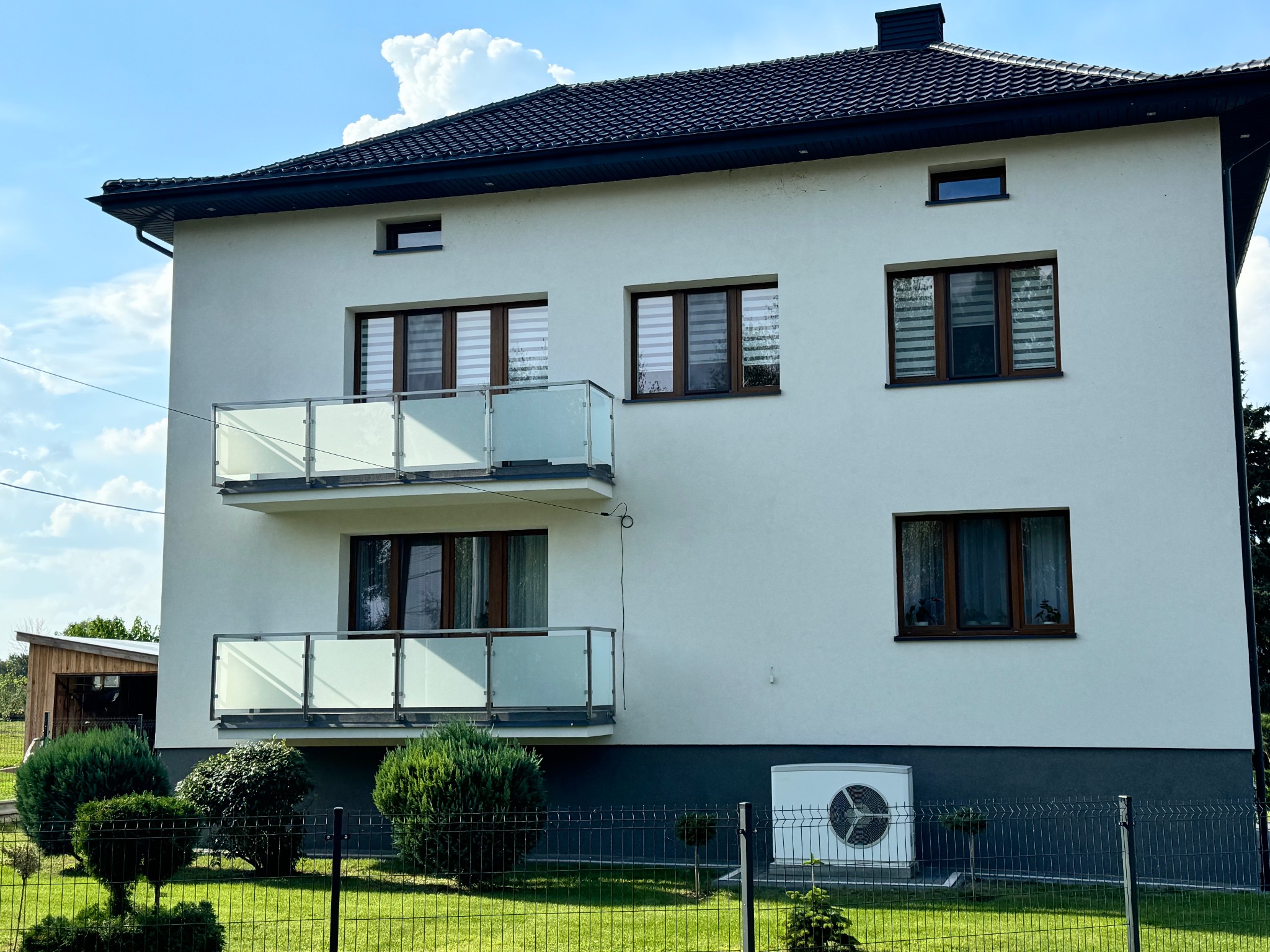 Nowoczesny dom z białym tynkiem i dwoma balkonami z szklanymi balustradami. Widoczne okna z roletami, zadbany trawnik i metalowe ogrodzenie.