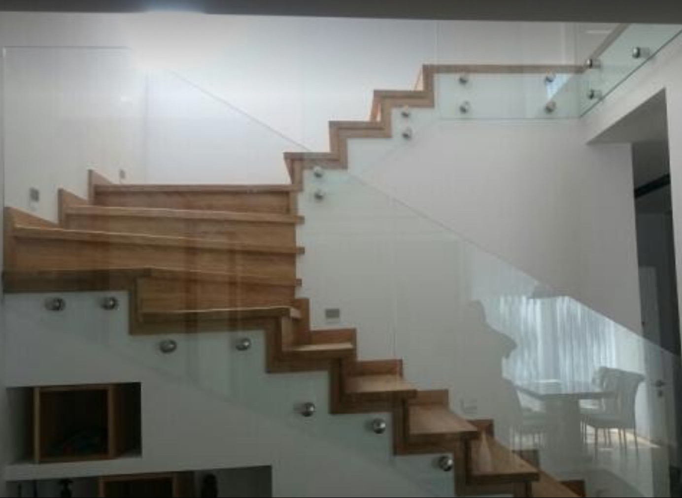 Schody drewniane z balustradą szklaną, mocowaną na metalowych uchwytach. Nowoczesny design, białe ściany, widok na jadalnię w tle. Eleganckie wykończenie wnętrza.