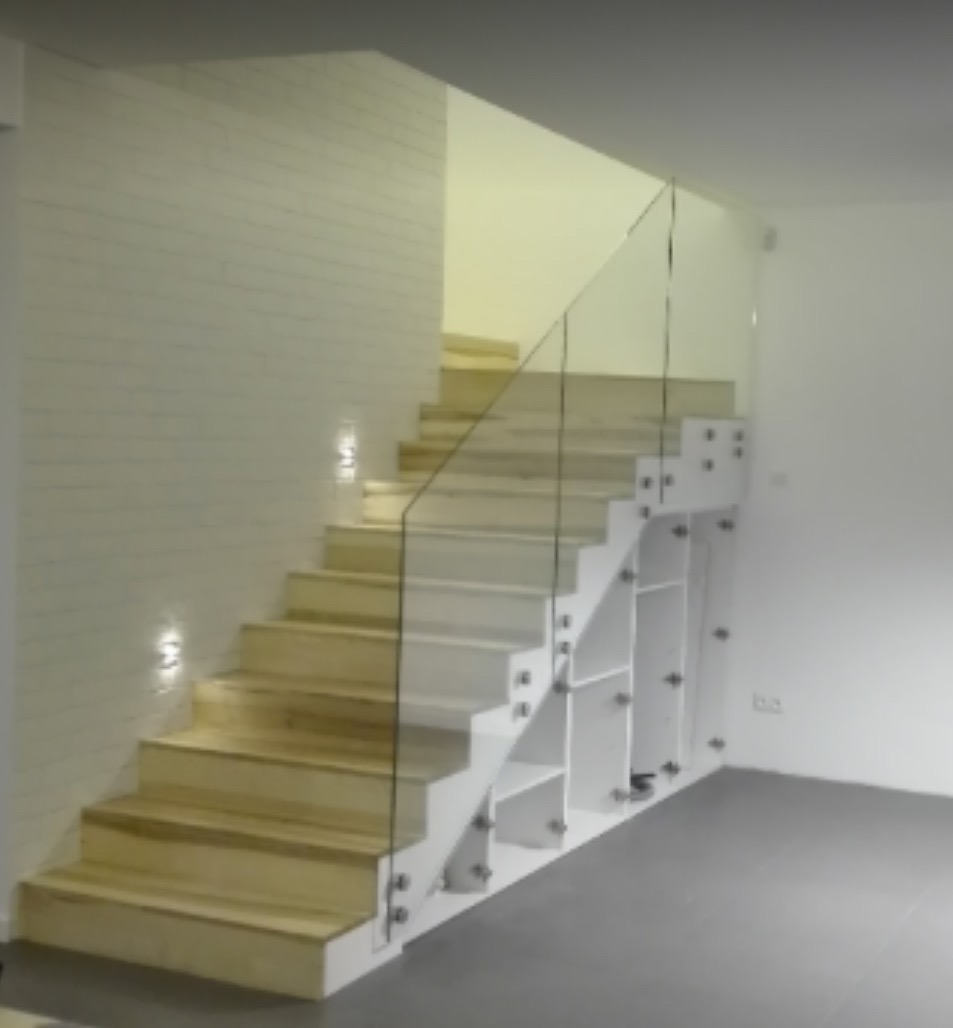 Jasne drewniane schody z przezroczystą szklaną balustradą, zamocowaną metalowymi uchwytami. Pod schodami białe półki. Ściana wykończona w cegiełkę.