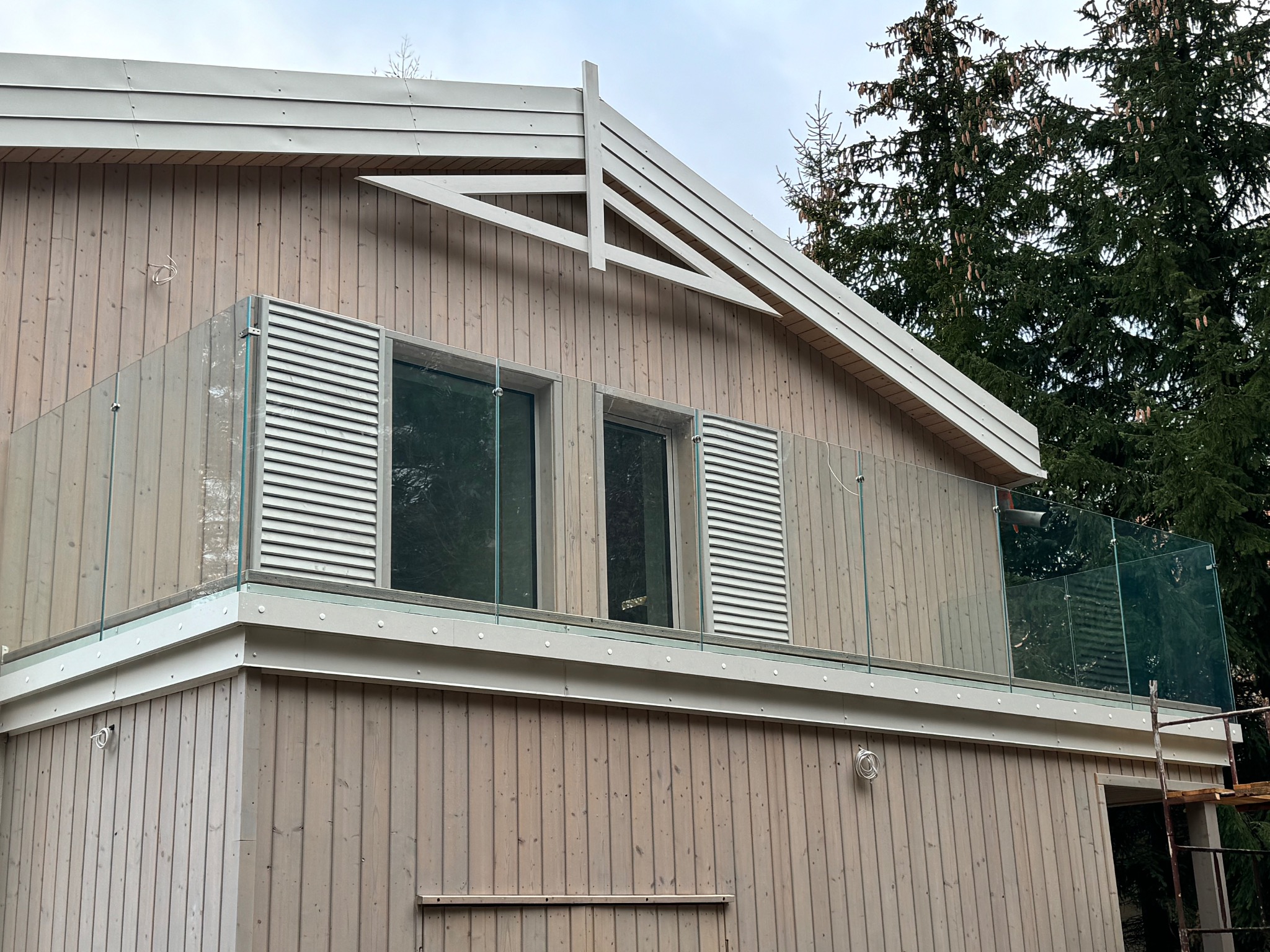 Nowoczesny balkon z balustradą szklaną, drewniane wykończenie elewacji, widok z dołu. Balustrada z elementami metalowymi i szkłem. Tło stanowią drzewa.