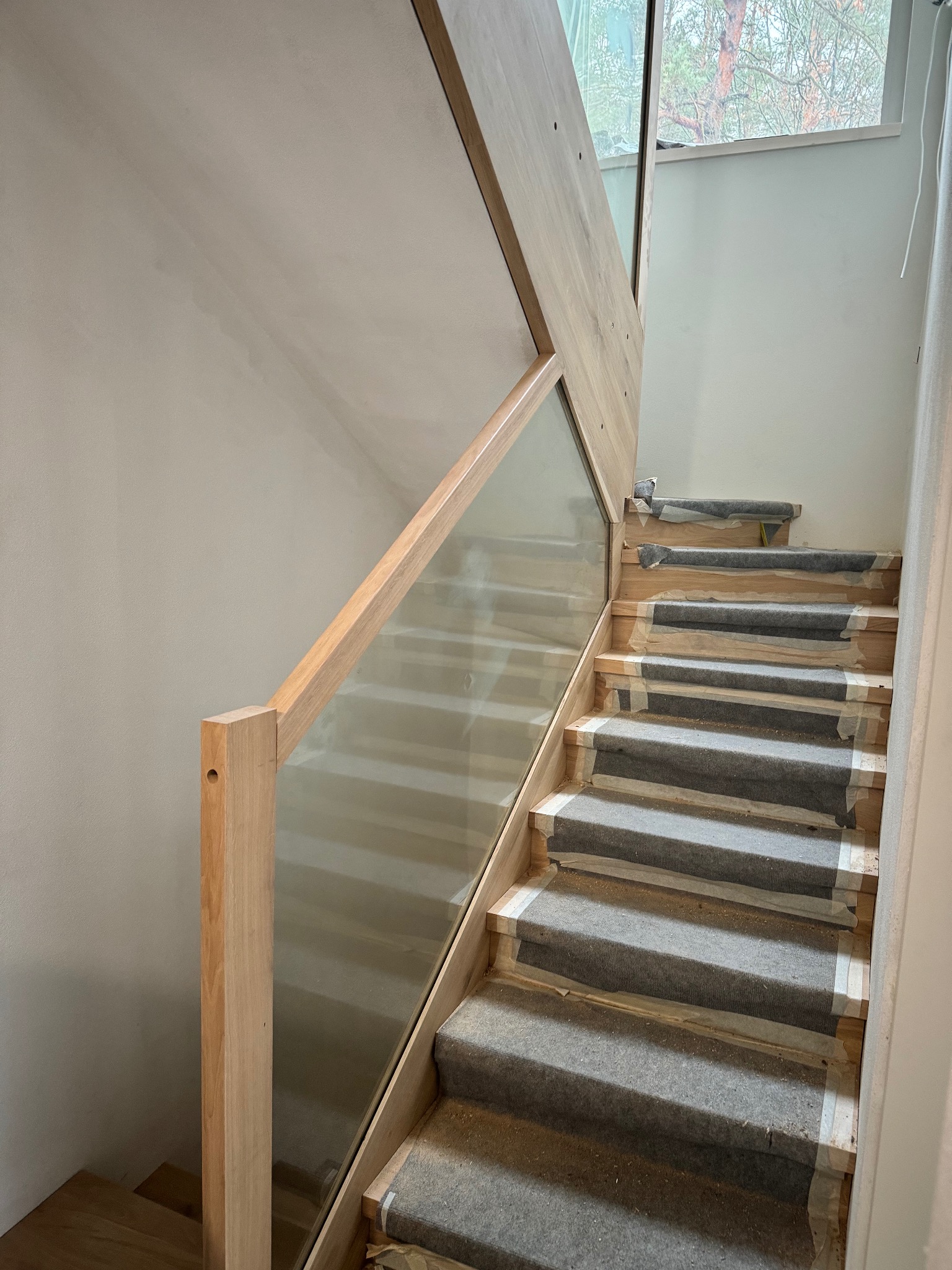 Nowoczesne schody z drewnianymi stopniami, zabezpieczone folią, z szklaną balustradą i drewnianą poręczą. Widok z dołu, jasne wnętrze.