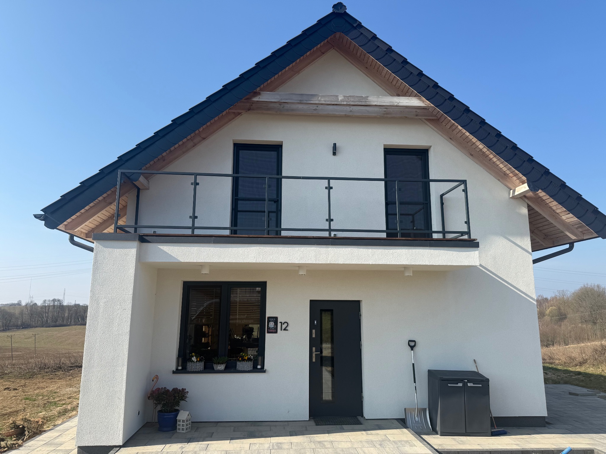 Biały dom z czarnym dachem i balkonem z nowoczesną balustradą ze szkła i metalu. Widoczne okna z czarnymi ramami, numer domu '12' i elementy dekoracyjne.