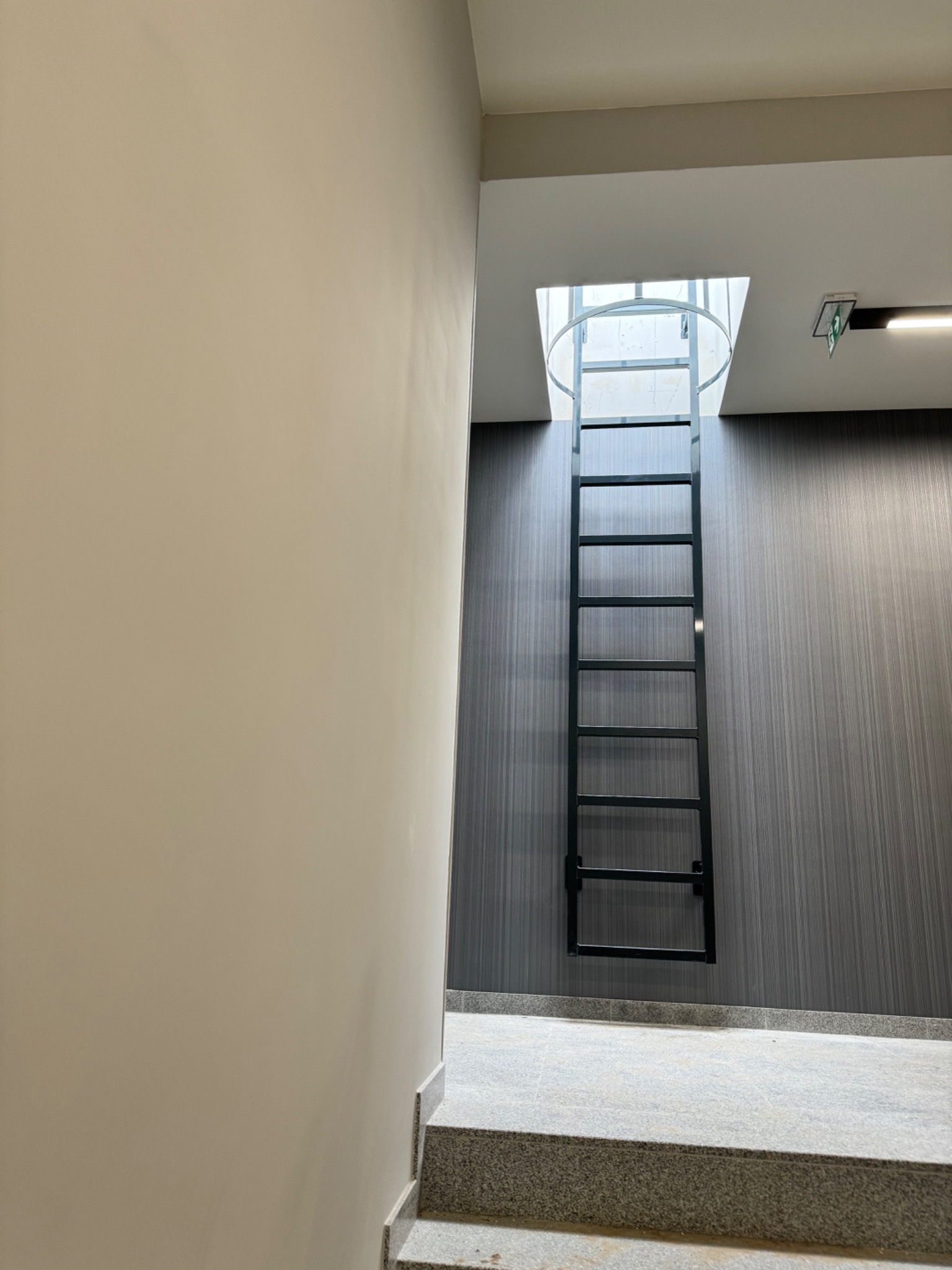 Metalowa drabina ewakuacyjna, przytwierdzona do ściany o prążkowanej strukturze, prowadząca do otworu w suficie z widocznym oświetleniem naturalnym.
