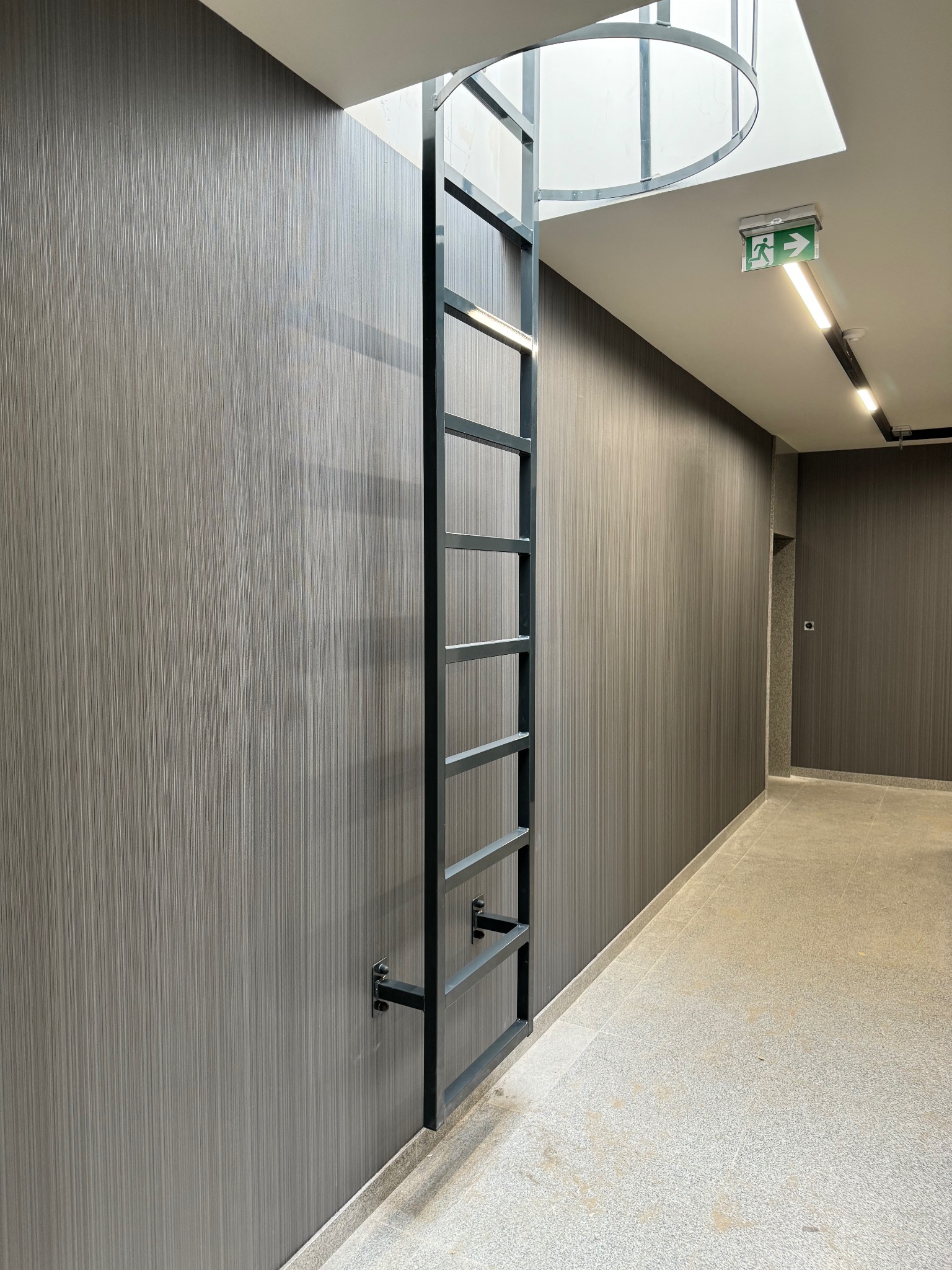 Metalowa drabina przymocowana do ściany w korytarzu, prowadząca do okrągłego otworu w suficie. Widoczny znak ewakuacyjny i oświetlenie.