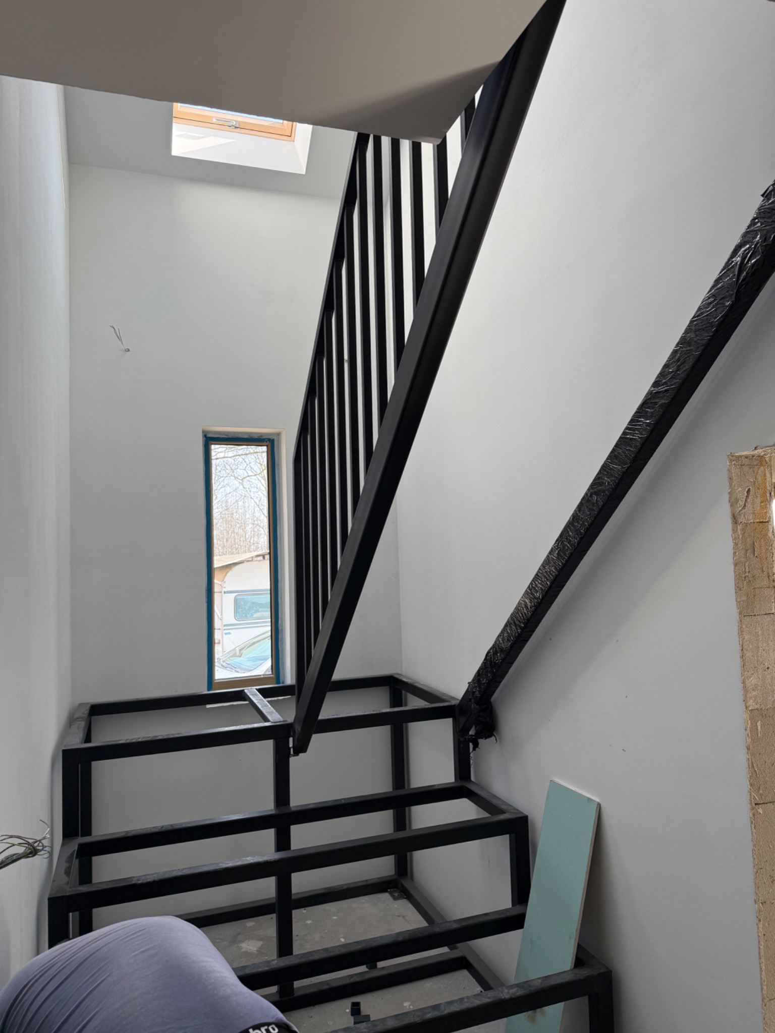 Wnętrze z metalową konstrukcją schodów z czarną balustradą. Widoczne okno i fragment osoby. Konstrukcja w trakcie montażu, zabezpieczona folią w górnej części.