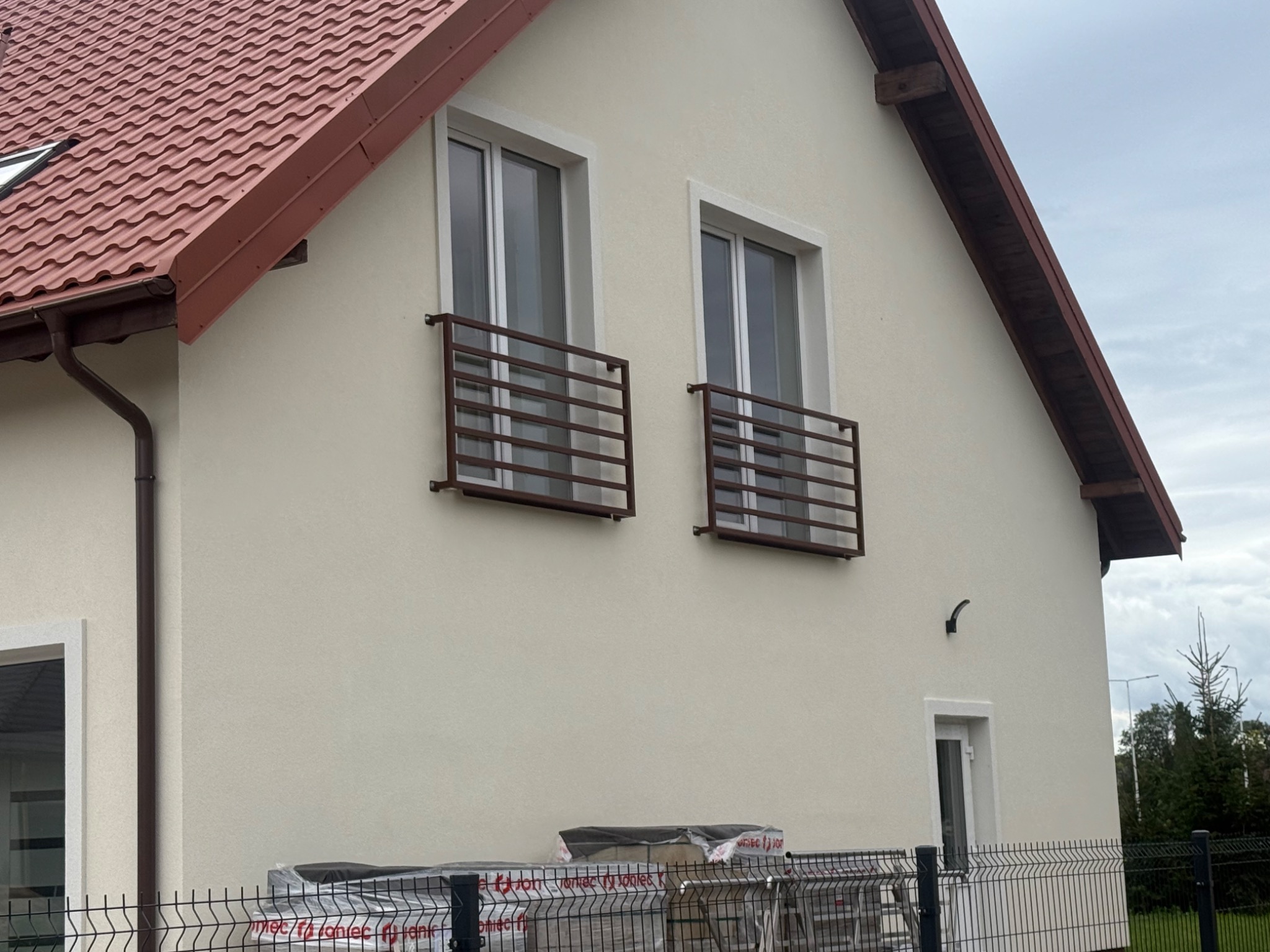 Dwa metalowe balkony z poziomymi prętami, zamontowane na elewacji domu z czerwoną dachówką. Widoczne okna, fragment ogrodzenia i materiały budowlane.