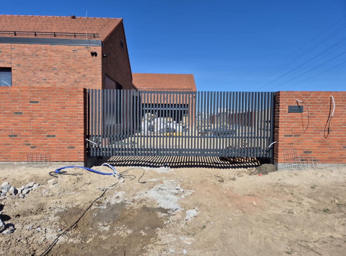 Nowoczesna brama wjazdowa z pionowych, szarych profili stalowych, wkomponowana w mur z czerwonej cegły na tle domu z czerwoną dachówką w słoneczny dzień.