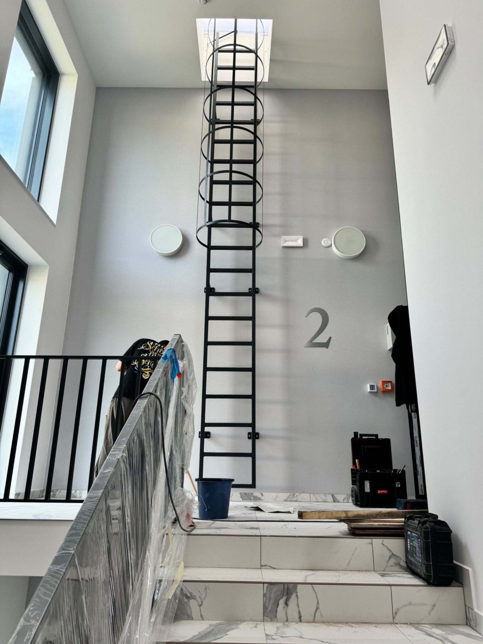 Wnętrze z metalową drabiną z obręczami ochronnymi prowadzącą do świetlika. Na schodach narzędzia i zabezpieczona folią balustrada. Widoczny fragment barierki i numer '2' na ścianie.