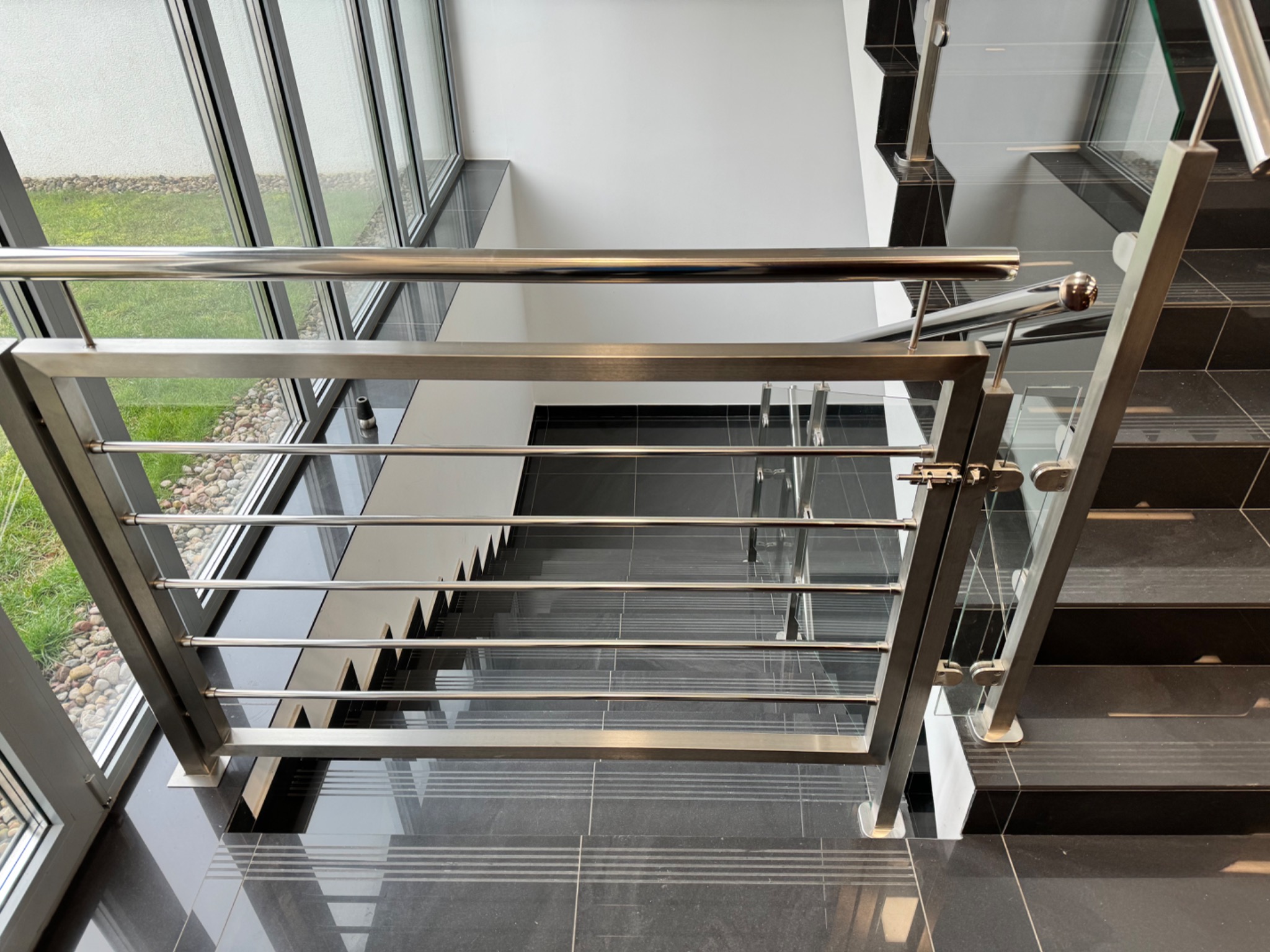 Nowoczesne schody z metalową balustradą i szklanymi elementami, widok z góry. Czarno-białe płytki podłogowe, duże okna z widokiem na zieleń.