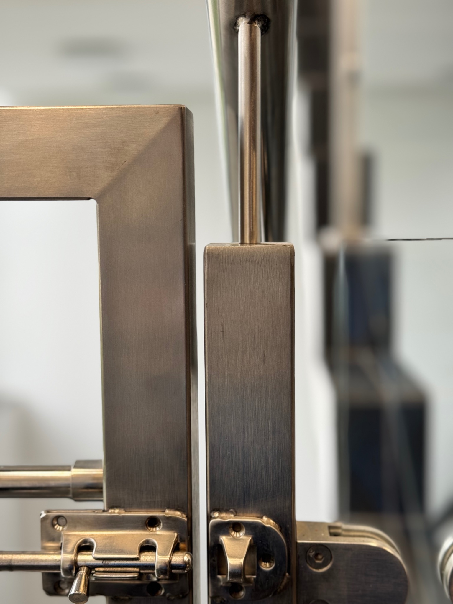 Szczegółowe ujęcie zamknięcia metalowej bramki z elementami szklanymi, widoczne zawiasy i zasuwa, szczotkowana stal nierdzewna, minimalistyczny design.