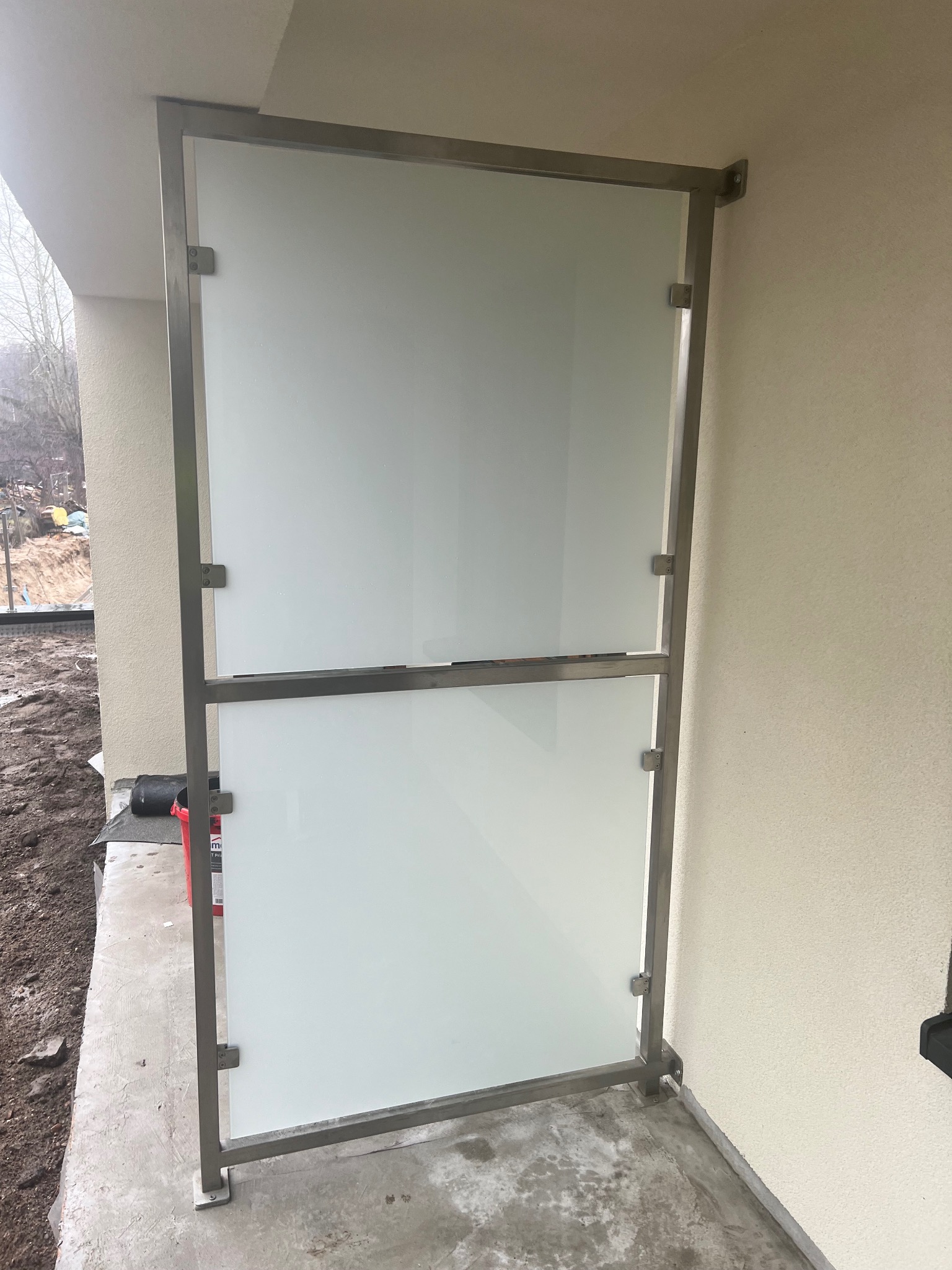 Szklana balustrada balkonowa z metalową ramą, zamontowana na betonowej posadzce. Widoczne mocowania i fragment elewacji budynku w neutralnym odcieniu.