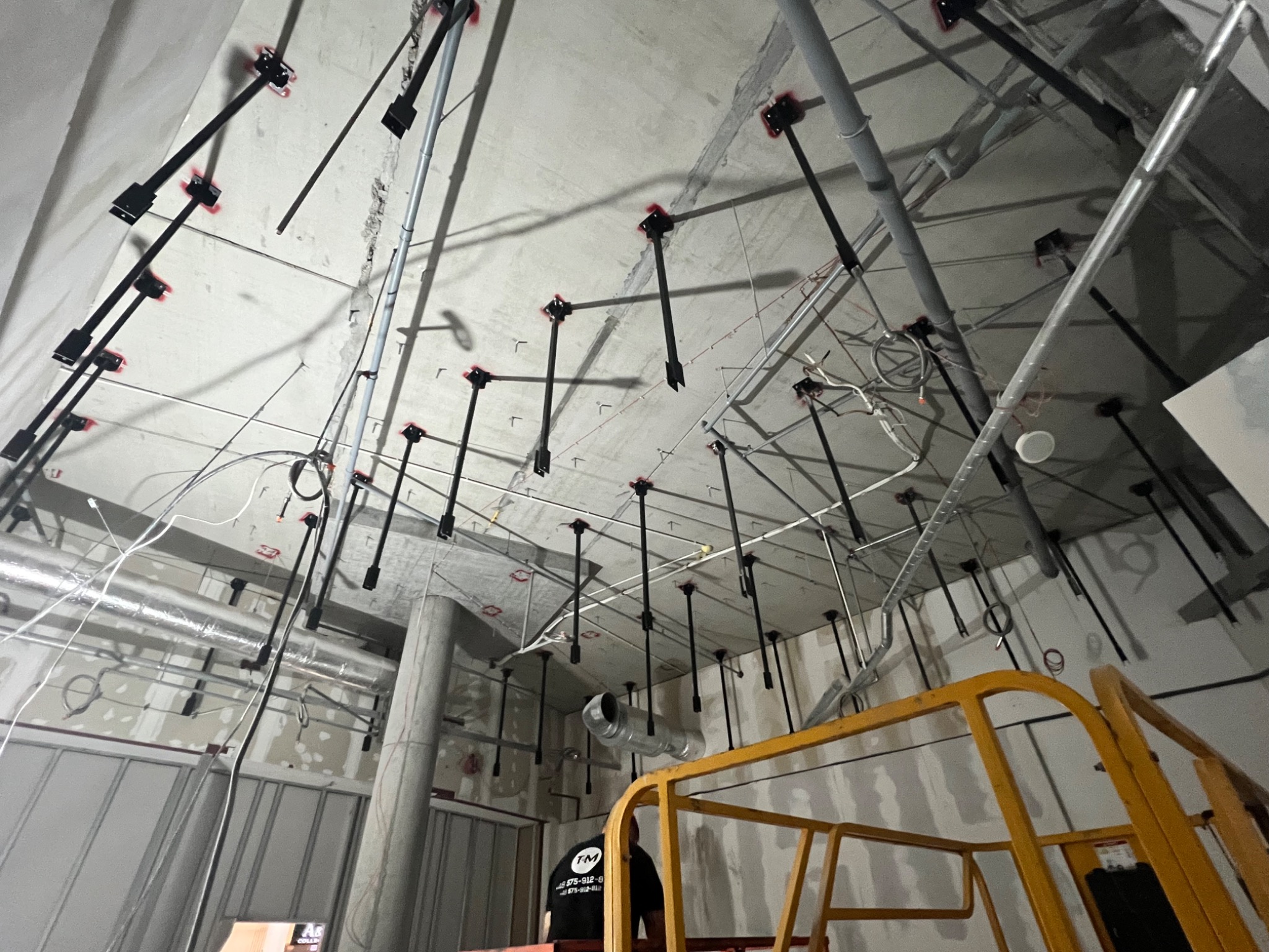 Montaż instalacji podwieszanej pod stropem, widoczne elementy mocujące i rury, w tle fragment podnośnika. Wnętrze w stanie surowym.