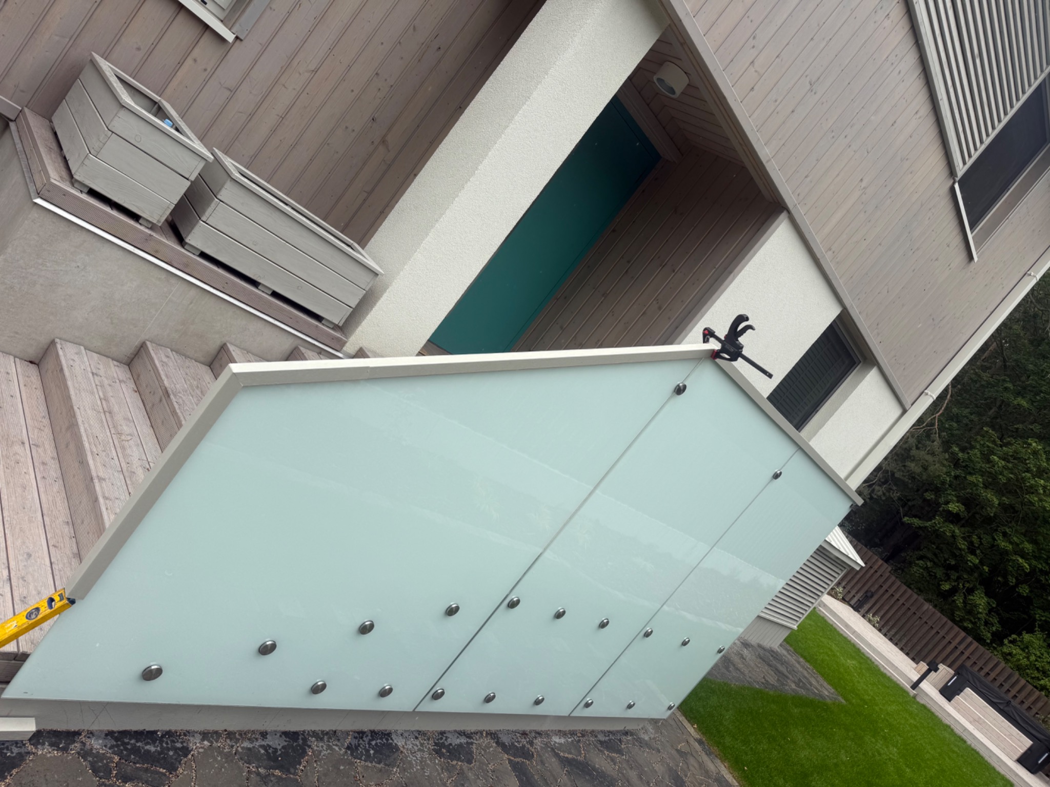 Nowoczesna balustrada szklana z mocowaniami punktowymi, widoczna z boku, z poziomnicą i ściskiem stolarskim. Tło: drewniana elewacja domu i trawnik.