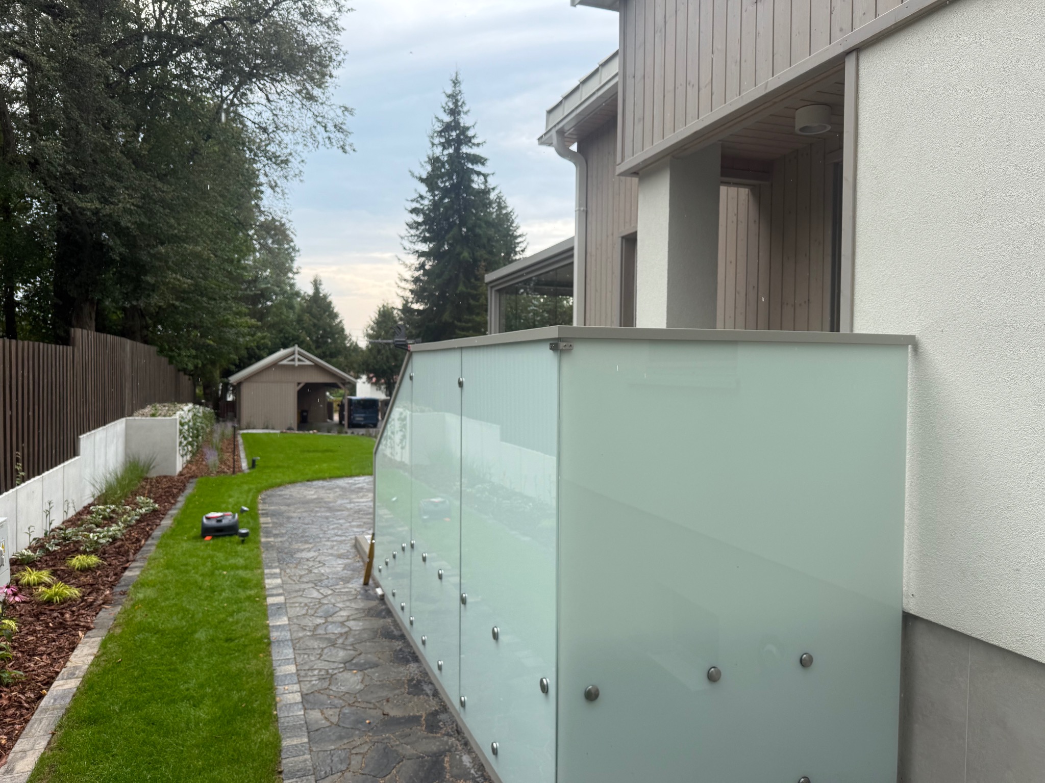 Nowoczesna zabudowa balkonu z mlecznego szkła, mocowana na metalowych elementach, z widokiem na zadbany ogród i dom w tle. Minimalistyczny design.