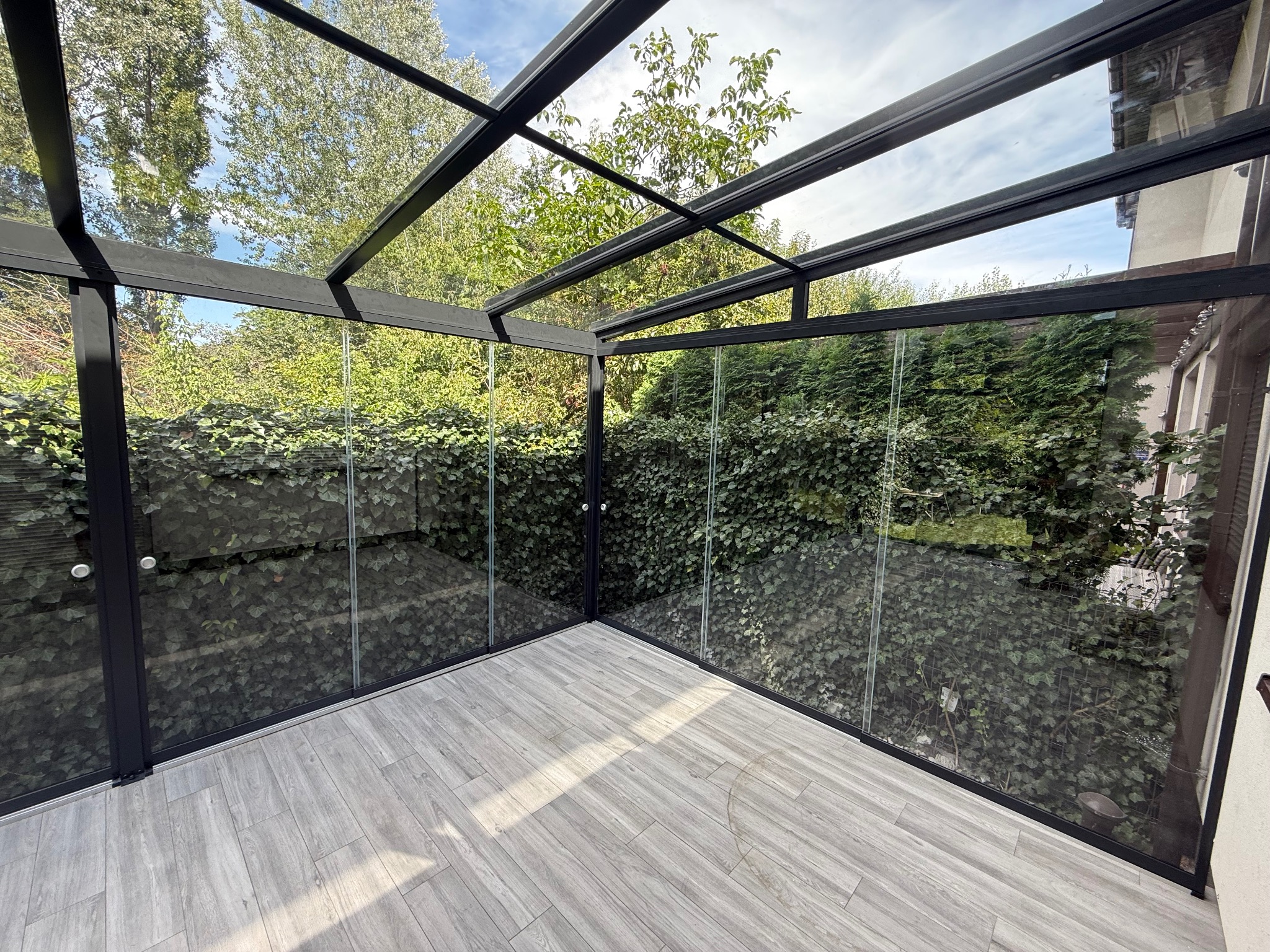 Nowoczesna zabudowa balkonu z czarną ramą i szklanymi ścianami, z widokiem na zielony ogród. Podłoga wyłożona panelami w odcieniach szarości.