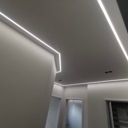 Staszel Bau - Mateusz Staszel - Nowoczesne oświetlenie LED wpuszczone w sufit, tworzące geometryczne linie światła w jasnym wnętrzu z widocznymi drzwiami.