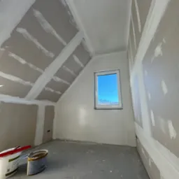 Pokój na poddaszu w trakcie prac wykończeniowych: ściany z płyt gipsowo-kartonowych ze szpachlowanymi łączeniami, widoczne dwa wiadra z masą szpachlową i szpachelka, okno z widokiem na błękitne niebo.
