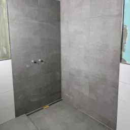 Szare, matowe płytki ceramiczne ułożone w kabinie prysznicowej z odpływem liniowym, widoczne białe rurki wystające ze ściany oraz fragmenty niezakończonych prac wykończeniowych na ścianach.